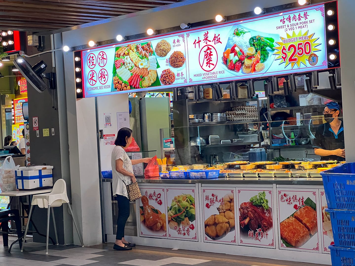 Woman at food stall with lighted menu displaying meal options, including "Economic Bee Hoon" and a "$2.50" budget meal.